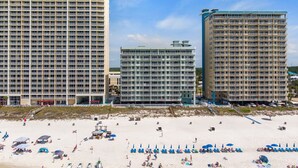 Un hotel frente a la playa con varios edificios altos, una playa de arena con sombrillas y tumbonas, y un cielo azul despejado.