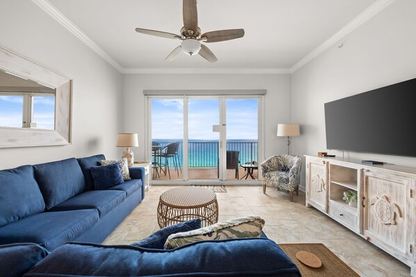 A living room with a blue sofa, a ceiling fan, a TV, and a balcony with a view of the ocean. - BEACH FRONT RESORT!! SLEEPS UP TO 9! BEACH SERVICE WITH 2 CHAIRS AND UMBRELLA INCLUDED! (Panama City Beach)