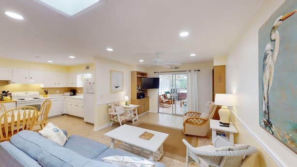 A spacious living area with a sofa, coffee table, and wicker chairs. A kitchen with white cabinets and a dining area with a table and chairs are visible.