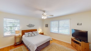 A bedroom with a bed, a ceiling fan, a TV, and two windows with blinds.