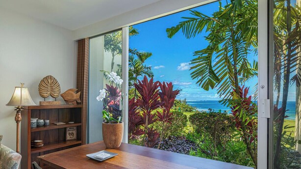 A dining area with a wooden table, chairs, a lamp, and a view of the ocean through large windows. - PUU POA #110 GORGEOUS OCEAN VIEWS AIR CONDITIONED Condo! (Princeville)