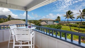 A balcony with white chairs and a table, overlooking a landscaped area with palm trees and a building in the background. - Poipu Sands 226  By Suite Paradise (Koloa)