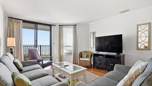 En moderne stue med grå sofaer, et sofabord i glas, et fjernsyn og en balkon med gardiner.