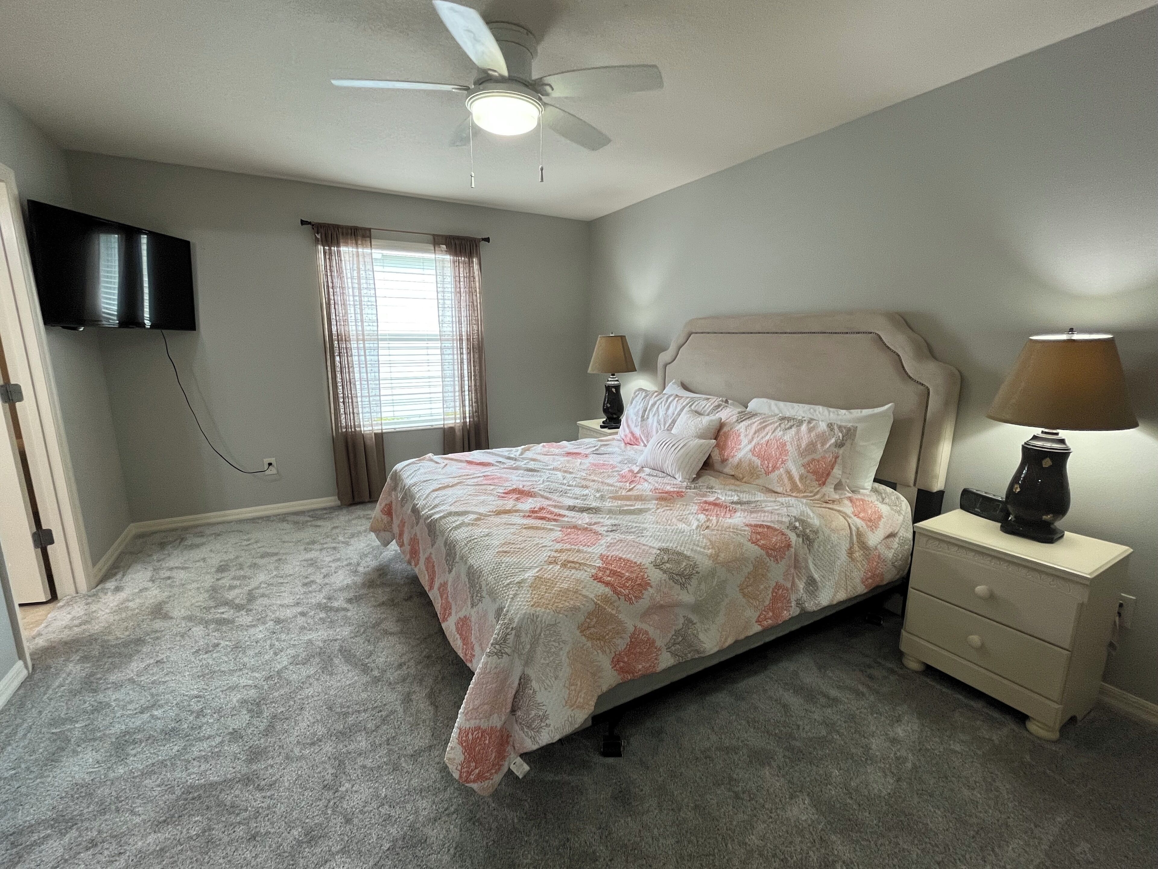 Une chambre à coucher comprenant un lit, des tables de chevet, un ventilateur de plafond, un téléviseur et une fenêtre avec des rideaux.