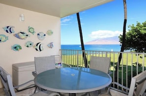 A balcony with a glass table and chairs, a white wall with fish decals, and a view of the ocean. - Royal Mauian #202 - Oceanview Gem Steps Away from Dining and Shopping! (Kihei)