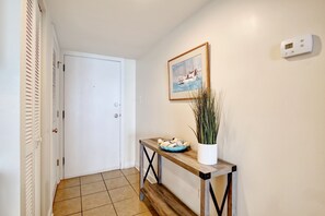 Un pasillo con una puerta blanca, un cuadro de veleros, un termostato digital, una consola de madera con un cuenco azul y una planta en maceta.
