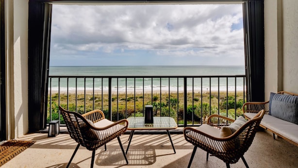 Ein Balkon mit Korbstühlen, einem Glastisch und Blick auf Strand und Meer.