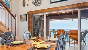 A dining area with a wooden table set for four, a staircase, and a view of the sea through a glass door. - Aloha Condos, Kanaloa at Kona, Condo 2004, Oceanfront, AC (Kailua Kona)