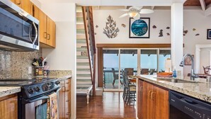 A kitchen with wooden cabinets, a stainless steel oven, a microwave, and a dishwasher. There is a dining area with a table and chairs, and a view of the ocean through large windows. - Aloha Condos, Kanaloa at Kona, Condo 2004, Oceanfront, AC (Kailua Kona)