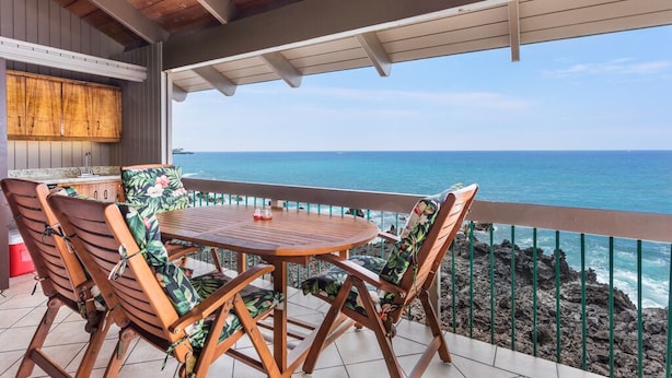 A covered outdoor seating area with a wooden table and chairs, a kitchen area with cabinets, and a view of the ocean. - Aloha Condos, Kanaloa at Kona, Condo 2004, Oceanfront, AC (Kailua Kona)