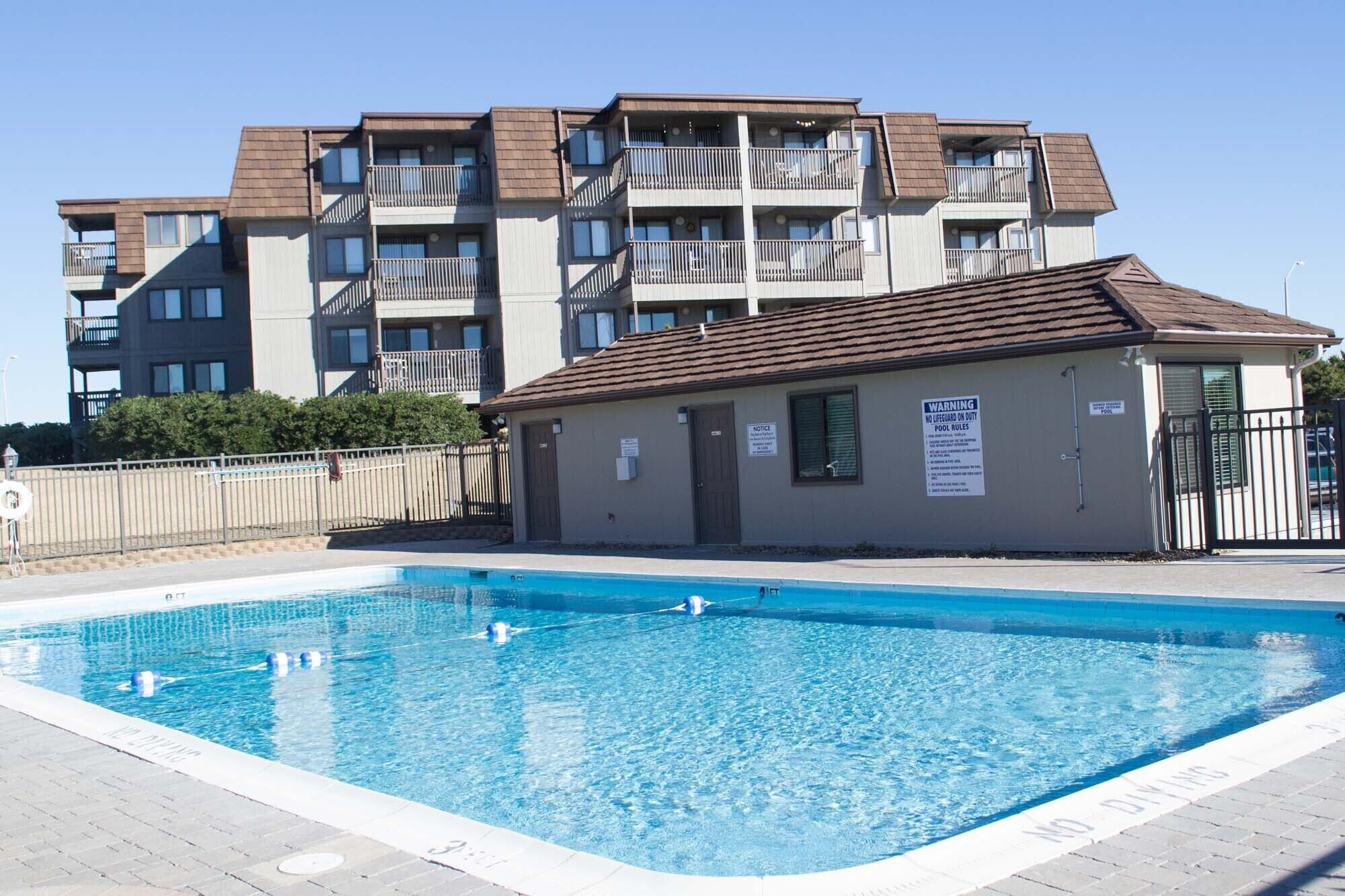 En swimmingpool med en klar, blå vandoverflade, omgivet af en flisebelagt terrasse. I baggrunden ses en fleretasagers boligbygning med altaner.