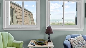 A room with two windows overlooking a beach, a green armchair, a table with a lamp, and a bowl.