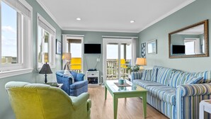 A living room with a green armchair, a blue sofa, and a striped sofa. There is a table with a lamp, a TV mounted on the wall, and a window with blinds.