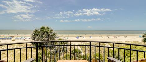 A balcony with wooden chairs overlooking a beach with umbrellas and people.