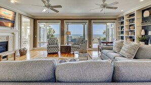 A spacious living room with a fireplace, two ceiling fans, and a view of the ocean through large windows.