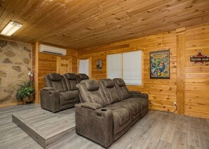 A wooden cabin interior with a stone wall, two reclining sofas, a stone fireplace, and a mounted air conditioning unit. - Free Nights! Unmatched Long Range Views, Hot Tub, Game room. Booking Summer now! (Pigeon Forge)