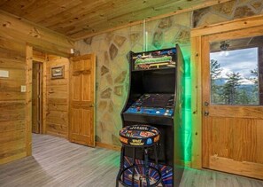 Um quarto de madeira com um jogo de fliperama com tema espacial, uma porta e uma janela com vista.