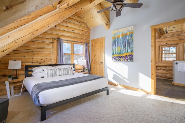 A log cabin bedroom with a bed, a ceiling fan, a painting, and a wooden door.