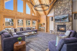 A log cabin interior with a stone fireplace, large windows, and a chandelier.