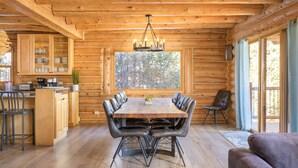 Interior de una cabaña de madera con una mesa de comedor, sillas, una araña y un ventanal grande.