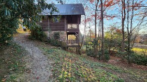 A rustic wooden cabin with a porch, surrounded by a forested area with a gravel path leading to it.