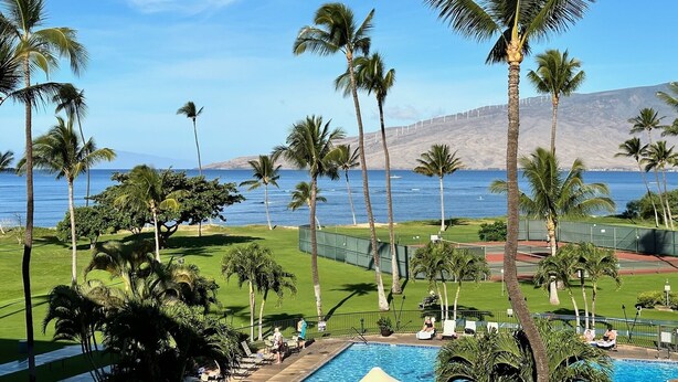 A pool area with palm trees, a view of the ocean, and a hilly landscape in the background. - Maui Sunset Vacation Rentals B415 (Kihei)