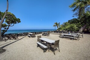 An outdoor seating area with tables and chairs, overlooking a beach with clear blue water. - 🏖️AC | Ocean View | Concierge Included (Kailua Kona)