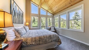 Un dormitorio con una cama grande, varios ventanales, techo de madera y vistas a árboles.