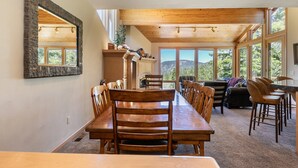 Un comedor amplio con una larga mesa de madera y sillas, ventanas grandes que ofrecen vistas al exterior y un alto techo con vigas de madera.