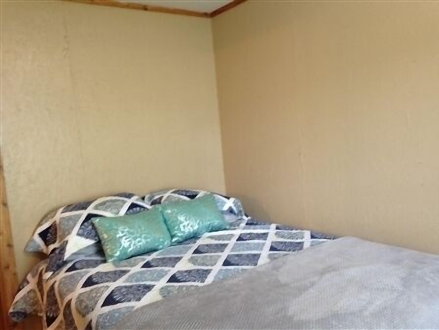 A small, single-bed room with a patterned bedspread and two pillows. The room has plain walls and a wooden ceiling.