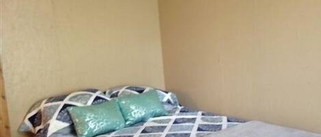 A small, single-bed room with a patterned bedspread and two pillows. The room has plain walls and a wooden ceiling.