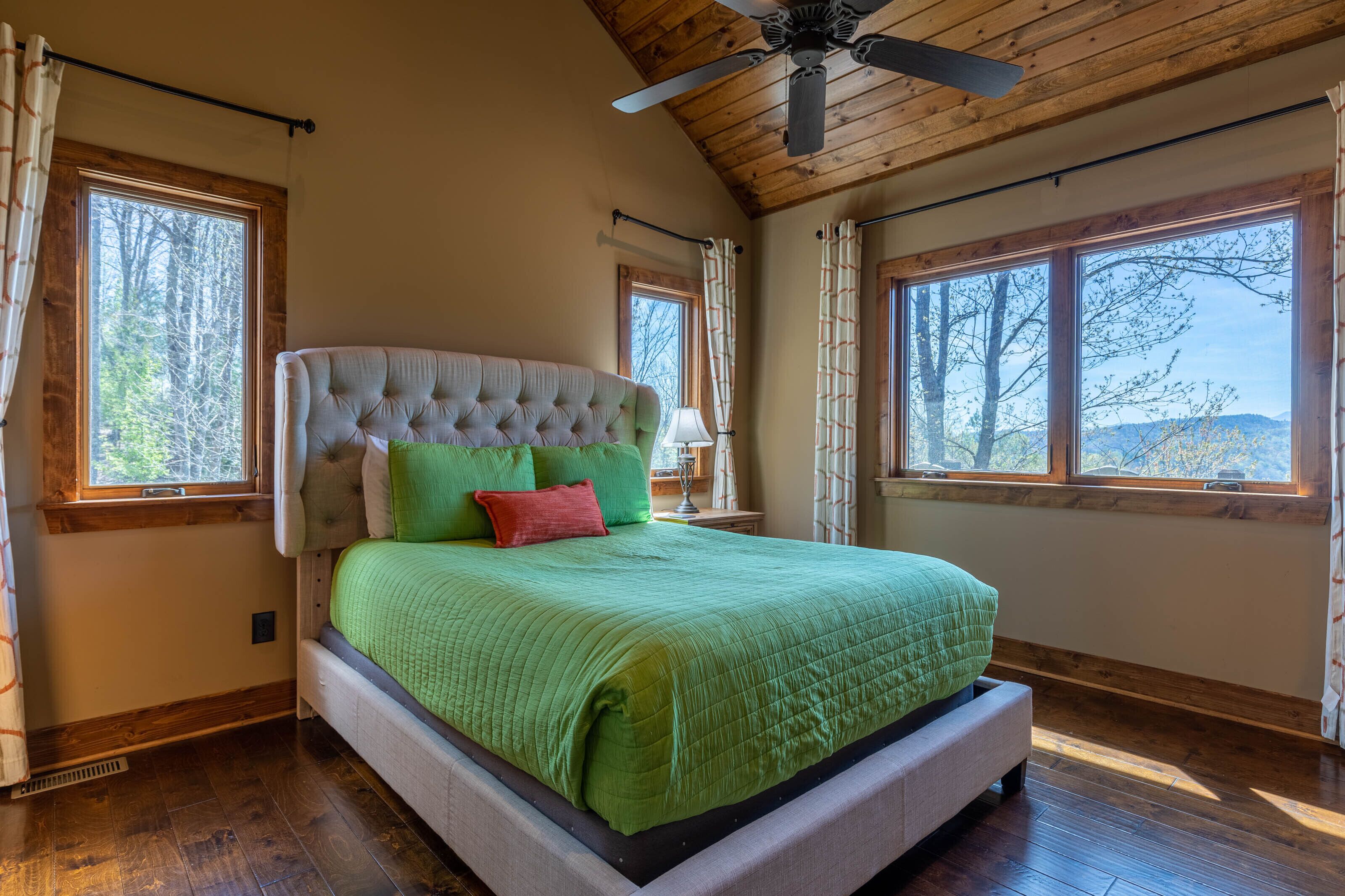 Una camera da letto con un letto grande, pavimento in legno e diverse finestre che offrono vista su alberi e colline.