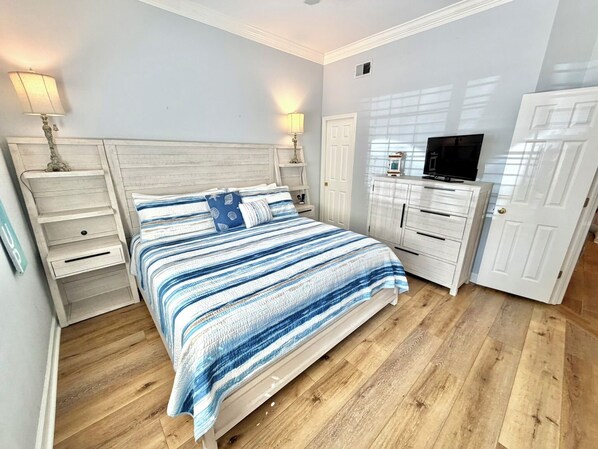 Un dormitorio con cama, mesitas de noche, cómoda, televisor y puerta.