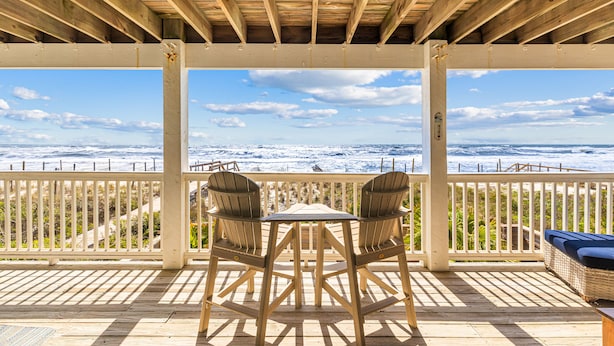 Um deck de madeira com mesa e cadeiras, com vista para uma praia, o oceano e o céu.