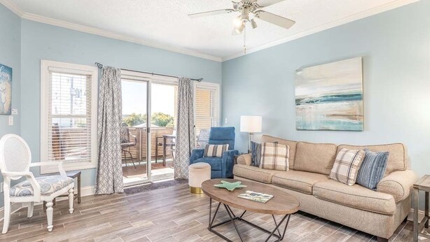Un salon avec un canapé beige, une table basse, un fauteuil bleu et une chaise blanche. On y trouve également un ventilateur de plafond, une fenêtre avec des rideaux et un tableau accroché au mur.
