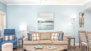 Un salon avec un canapé d’angle beige, une table basse ronde, un fauteuil bleu et une table à manger en bois.