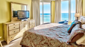 Une chambre d’hôtel avec un grand lit, un téléviseur posé sur une commode en bois et une vue sur l’océan par de grandes fenêtres.
