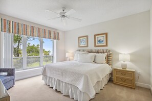 Ein Schlafzimmer mit einem großen Bett, einem Schreibtisch und einem Stuhl. An der Wand hängen zwei gerahmte Bilder, ein Fenster mit Vorhängen und ein Deckenventilator.