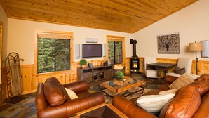 Ein gemütliches Wohnzimmer mit Ledersofas, einem Kamin, einem Fernseher und großen Fenstern mit Blick auf einen Wald.