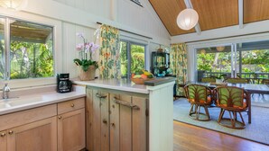 A kitchen with wooden cabinets, a coffee maker, and a fruit bowl. There is a dining area with wicker chairs and a table. - Hale Pua Kai, Comfortable Hawaiian Style Home (Haena)