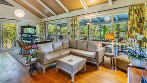 A spacious living area with a large sectional sofa, a coffee table, wicker furniture, and large windows offering a view of lush greenery. - Hale Pua Kai, Comfortable Hawaiian Style Home (Haena)