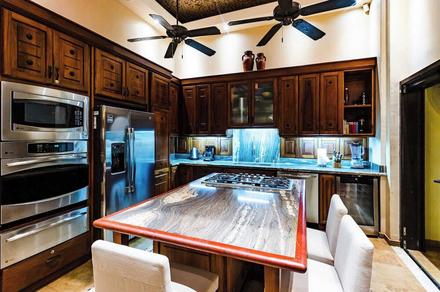 Een moderne keuken met houten kastjes, roestvrijstalen apparatuur, een marmeren aanrechtblad en een plafondventilator.