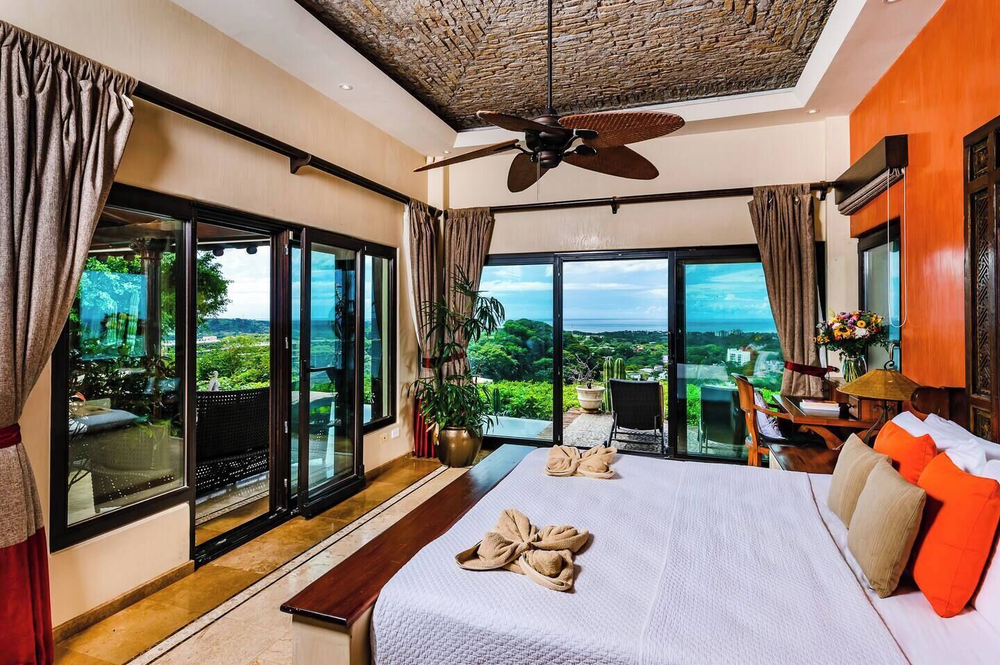 Un dormitorio amplio con una cama grande, un ventilador de techo y vistas a la exuberante vegetación y un paisaje urbano lejano a través de amplias puertas correderas.