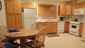 A compact kitchen with wooden cabinets, a white refrigerator, a microwave, a stove, and a sink. There is a dining table with chairs and a small rug on the floor. - Spacious 2 Bedroom Apartment above Vacation Home (Almont)