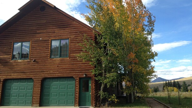 A wooden cabin with two green garage doors, a front porch, and a tree with autumn leaves. - Spacious 2 Bedroom Apartment above Vacation Home (Almont)