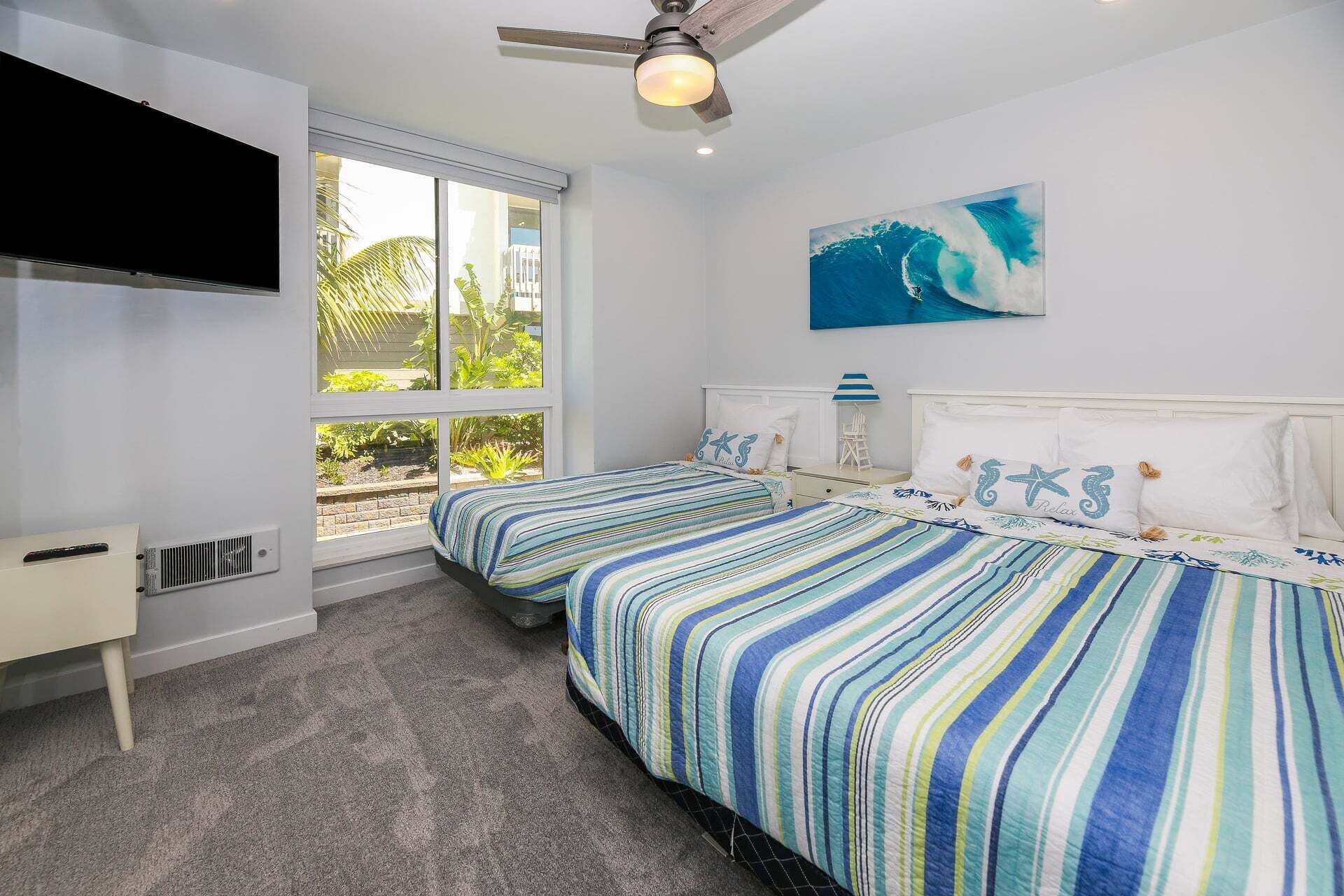 A bedroom with two beds, a ceiling fan, a TV, and a window with a view of greenery.
