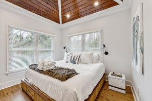 Quarto com teto de madeira, paredes brancas e uma cama grande com roupa de cama branca. Há duas janelas com persianas, uma mesinha de cabeceira e uma pequena mesa lateral.