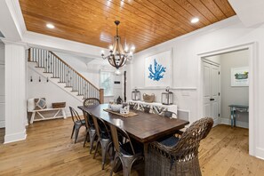 A dining area with a wooden table, metal chairs, a chandelier, and a staircase with white railings. - Luxury Vaycay! Private Pool, Billiards, Hot Tub, Putting Green - 2602 Hartnett (Isle of Palms)
