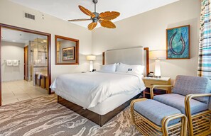 Une chambre d’hôtel avec un lit, une chaise, un bureau avec une lampe, un tableau et un ventilateur de plafond.