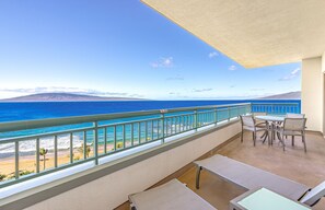Un balcon avec vue sur l’océan et une plage. Des tables et des chaises sont à disposition pour s’asseoir.